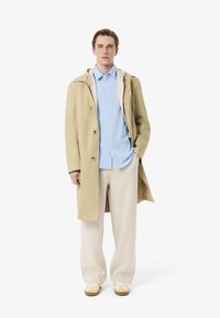 Impermeabile beige con cappuccio grigio chiaro, camicia blu e pantaloni beige. Le calzature sono sneaker gialle con accenti marroni.