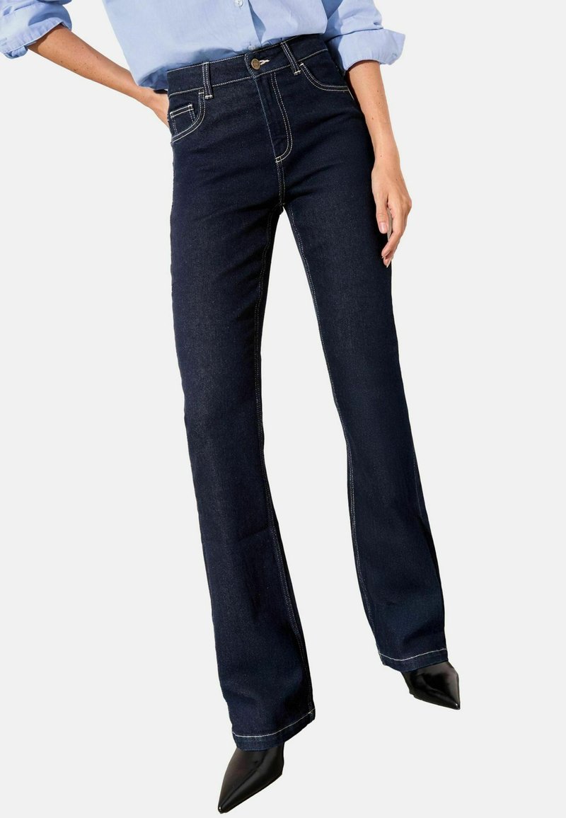 Jeans de denim oscuro de pierna acampanada con cintura alta, adornados con costuras de contraste y bolsillos frontales. Textura suave, combinados con tacones negros de punta.