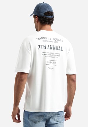 Man draagt een witte oversized t-shirt met de tekst "7th Annual Market Square" op de achterkant, lichtblauwe spijkerbroek en een blauwe baseballpet, kijkt weg.