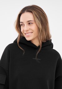 Felpa nera con un fit rilassato, dotata di cappuccio con cordino e un piccolo logo sul petto. Il tessuto sembra morbido, con una superficie liscia.