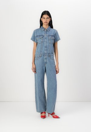 Denim jumpsuit with short sleeves, two front pockets, and a fitted waist. Light blue color and wide-leg pants. Paired with red high-heeled shoes.