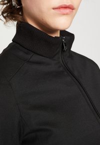 Svart zip-up jacka med ribbad krage. Den har ett slätt, texturerat tyg med sidopaneler och en metallic dragkedja.