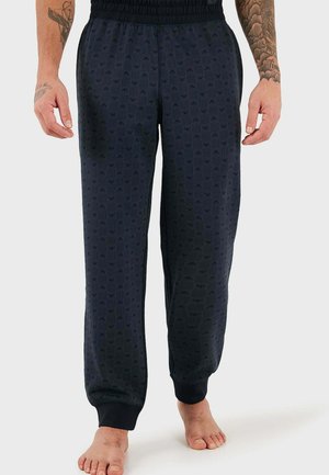 Hombre con pantalones jogger negros con estampado, cintura y puños elásticos, de pie descalzo contra un fondo claro y liso.