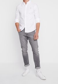Camisa blanca de botones con un pequeño logo, jeans de mezclilla gris y zapatillas blancas. El conjunto presenta un ajuste casual y puños enrollados.