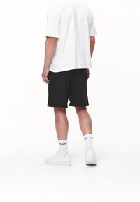 Osoba stojąca tyłem do widza, ubrana w białą koszulkę, czarne szorty, białe skarpety z tekstem oraz białe tenisówki na białym tle.
