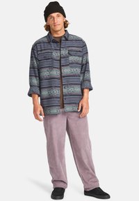 Chemise en flanelle multicolore avec un motif stylisé, fermeture à boutons, et deux poches poitrine, associée à un pantalon en velours côtelé violet clair et des chaussures noires.