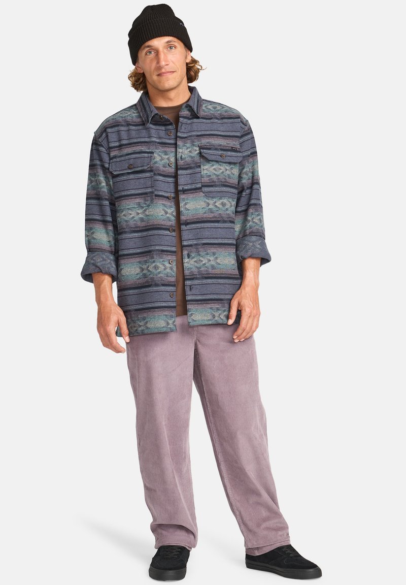 Chemise en flanelle multicolore avec un motif stylisé, fermeture à boutons, et deux poches poitrine, associée à un pantalon en velours côtelé violet clair et des chaussures noires.