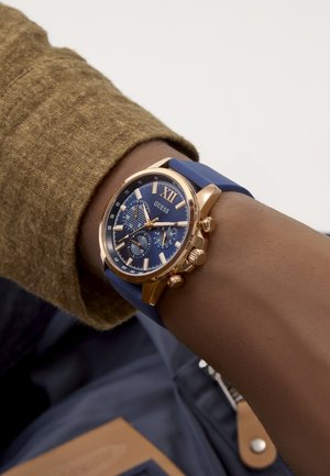 Polso con orologio cronografo Guess in oro e blu con cinturino in silicone blu e manica marrone dalla trama visibile.