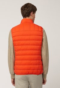 Chaleco acolchado naranja con cuello alto y acolchado horizontal. Llevado sobre una camiseta de manga larga beige, que muestra una textura interior suave.