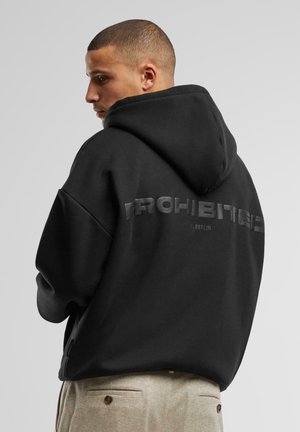 Man with short hair wearing a black hoodie with "PROHIBITED" text and beige pants, looking to the side against a plain background.
