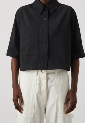 Person wearing a black short-sleeve button-up shirt with a chest pocket paired with high-waisted white pants with front pockets.