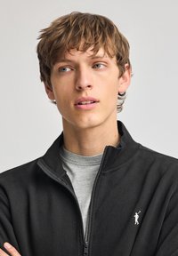 Svart zip-up jacka med hög krage, texturerat tyg, med en broderad logotyp på vänster sida. Grå skjorta under.