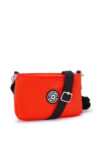 Borsa a tracolla rossa in nylon con chiusura a zip, tracolla nera regolabile e un patch rotondo con logo. Presenta un dettaglio in pom-pom testurizzato.