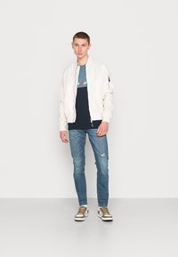 Casaco bomber branco, t-shirt azul-marinho com logótipo, jeans azul descolorido e sapatilhas castanhas com detalhes brancos. Design simples, visual casual.