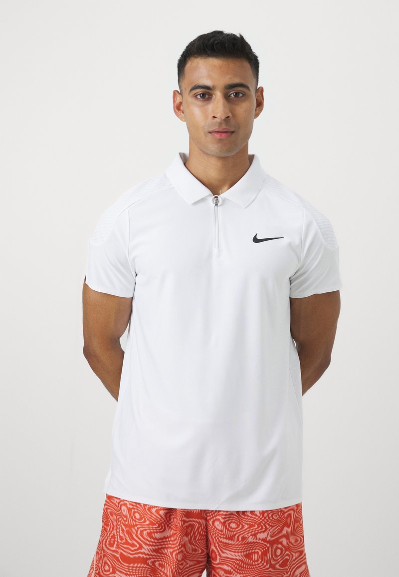 Biały polo z kołnierzykiem i zamkiem błyskawicznym do połowy, z czarnym logiem Nike na piersi. Noszone z wzorzystymi pomarańczowymi szortami. Gładka tekstura.