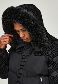 Čierna prešívaná bunda s vzorovanou hornou časťou, vybavená kapucňou z umelého kožišiny. Obsahuje zipsové vrecká a vystúpené logo na prednej strane.