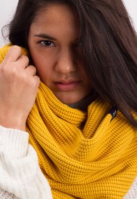 Knitted yellow infinity scarf draped around neck, featuring a textured pattern and subtle branding detail, paired with a light sweater.