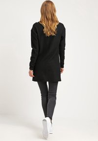 Tunic nera a maniche lunghe in maglia, texture morbida, lunghezza a metà coscia, accompagnata da pantaloni grigi slim e sneakers bianche. Vista posteriore.