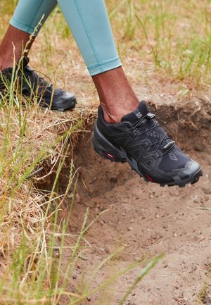 Personne portant des chaussures de trail noires et des leggings bleu clair descendant d'un talus de terre dans un terrain herbeux.