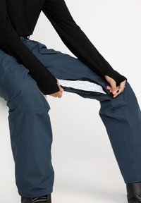 Blue insulated pants with a zippered opening and white mesh lining. They are paired with a black long-sleeve top and black boots.