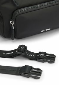 Borsa nera in nylon con una texture liscia, tasca con zip e spallacci regolabili con logo "HUGO" bianco e hardware in plastica.