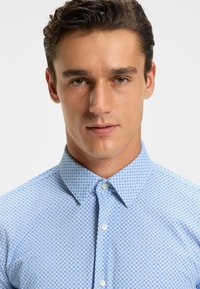 Vaaleansinisessä napitettavassa paidassa on tummansinisestä geometrisestä kuviosta, klassinen kaulus ja sileä kangastuntu.