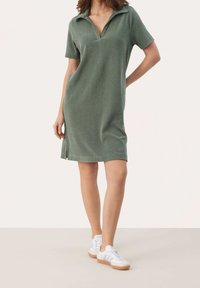Polo-tyylinen mekko teksturoidussa vihreässä kankaassa, lyhyillä hihoilla ja V-pääntiellä. Sisältää sivureikät ja on yhdistetty valkoisiin lenkkareihin.
