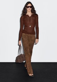 Hnědý žebrovaný top s dlouhým rukávem, hnědé semišové kalhoty a hnědá kabelka. Modelka má tmavé sluneční brýle a stojí na černé podlaze.