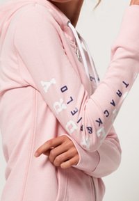 Sudadera de cremallera color rosa claro con cordones blancos; cuenta con letras azules a lo largo de las mangas. Material suave con puños acanalados.