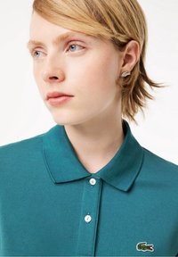 Ung person med kort, blont hår iført en teal polo-shirt med krokodillelogo og en sølvfarvet krokodilleformet ørering, der kigger til venstre.