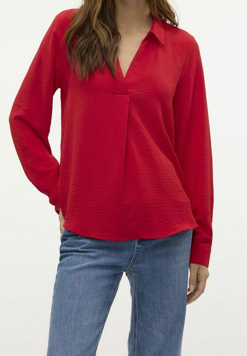Femme portant un chemisier rouge texturé à manches longues avec un col en V, associé à un jean en denim bleu, posant avec une main sur la hanche.