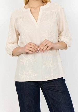 Femme portant un chemisier crème avec broderie florale et manches trois-quarts, associé à un jean bleu foncé, mains jointes à la taille.