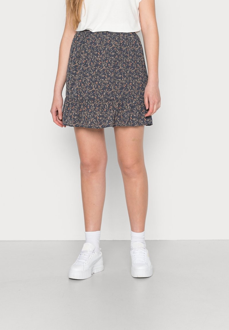 Femme portant une jupe bleu marine à motif floral, un haut blanc, des chaussettes blanches montantes et des baskets compensées blanches, debout devant un fond uni clair.