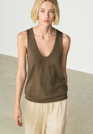 Donna con capelli corti biondi che indossa una canotta marrone senza maniche con scollo a V e pantaloni beige chiaro larghi, in piedi su un pavimento di cemento uniforme.