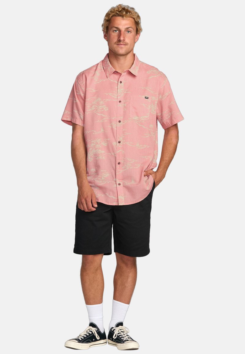 Kurzärmliges rosa Hemd mit hellbeigem Palmenmuster, Brusttasche, kombiniert mit schwarzen Shorts und schwarzen Turnschuhen.