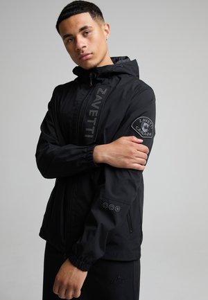 Giovane uomo con giacca nera Zavetti Canada con cappuccio, in piedi con le braccia incrociate davanti a uno sfondo grigio uniforme.