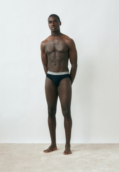 Μυώδης άνδρας όρθιος ξυπόλητος σε δάπεδο με χαλί μπροστά σε λευκό τοίχο, φορώντας μαύρα εσώρουχα με γκρι ζώνη στη μέση.