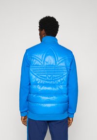 Homem a usar um casaco acolchoado azul brilhante com um grande logótipo em trevo cozido nas costas, combinado com calças azul-marinho com riscas laterais.