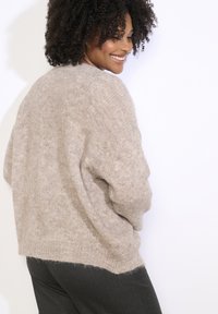 Bēšs adīts džemperis ar mīkstu, teksturētu apdari. Iespējas ar vaļīgu piegriezumu un ribotu apakšmalu, kombinēts ar tumšiem biksēm.