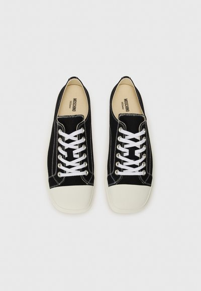 Svarta kanvas sneakers med vita snören, vit gummitå, och kontrasterande stygn. Interiören har "MOSCHINO"-logotyp.