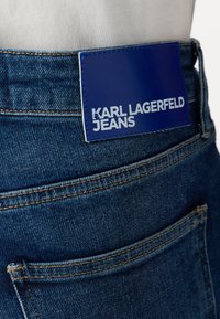 Jeans de mezclilla azul con un bolsillo trasero visible, que presenta una etiqueta de cuero azul con el logo "KARL LAGERFELD JEANS." Detalles cosidos que realzan la textura.