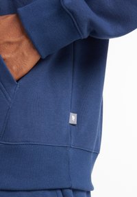 Mano in tasca del felpa blu New Balance, che mostra il polsino a coste e il piccolo logo sul lato della cucitura.