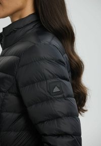 Sort pufjakke med standkrave og vandret quiltet tekstur. Har et struktureret sort mærke med et bjergdesign på ærmet.