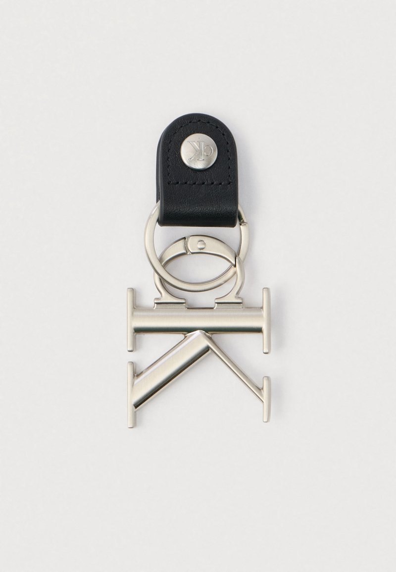 Srebrny metalowy breloczek z geometrycznymi kształtami przymocowany do czarnej skórzanej taśmy z metalowym guzikiem grawerowanym "ck" na białym tle.
