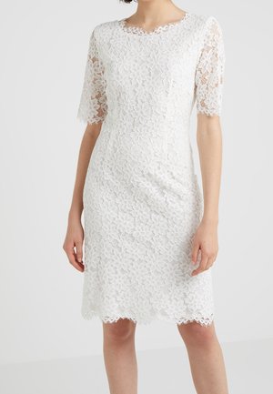 Femme portant une robe blanche en dentelle fleurie à longueur genou, avec des manches courtes et des bords festonnés aux manches et à l'ourlet.