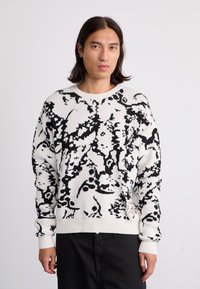 White knitted sweater featuring a bold black abstract pattern, round neckline, ribbed cuffs, and a small tag on the hem.