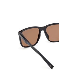Czarne okulary przeciwsłoneczne o smukłej, kątowej oprawie, brązowych szkłach przyciemnianych oraz cienkiej, teksturowanej czarnej zauszniku z detalem logo.