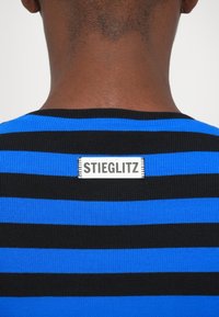 Gros plan sur le cou et le haut du dos d'une personne portant une chemise rayée bleue et noire avec une étiquette blanche portant la mention "STIEGLITZ."