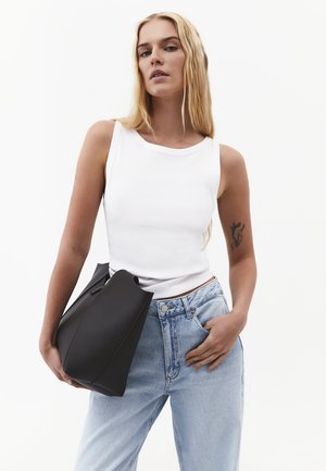 Mujer con camiseta blanca sin mangas y vaqueros azul claro sosteniendo una gran bolsa negra, con un tatuaje visible en su brazo izquierdo superior.