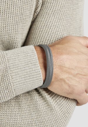 Handgelenk mit gewebtem grauem Lederarmband über einem Ärmel aus beigefarbenem Strickpullover, Hand ruht auf dem gegenüberliegenden Arm.